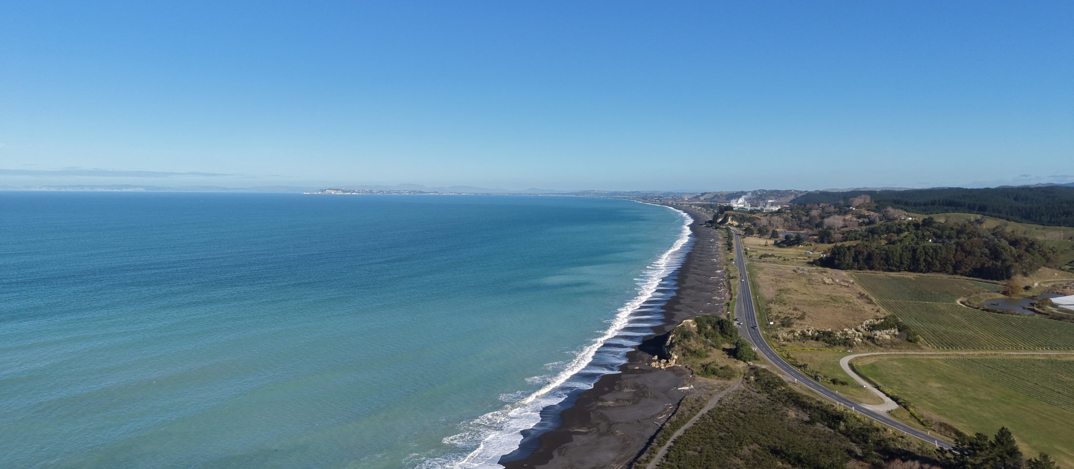 The Hawke's Bay coast from the air: surf on a black-sand beach, the highway and green paddocks beside it