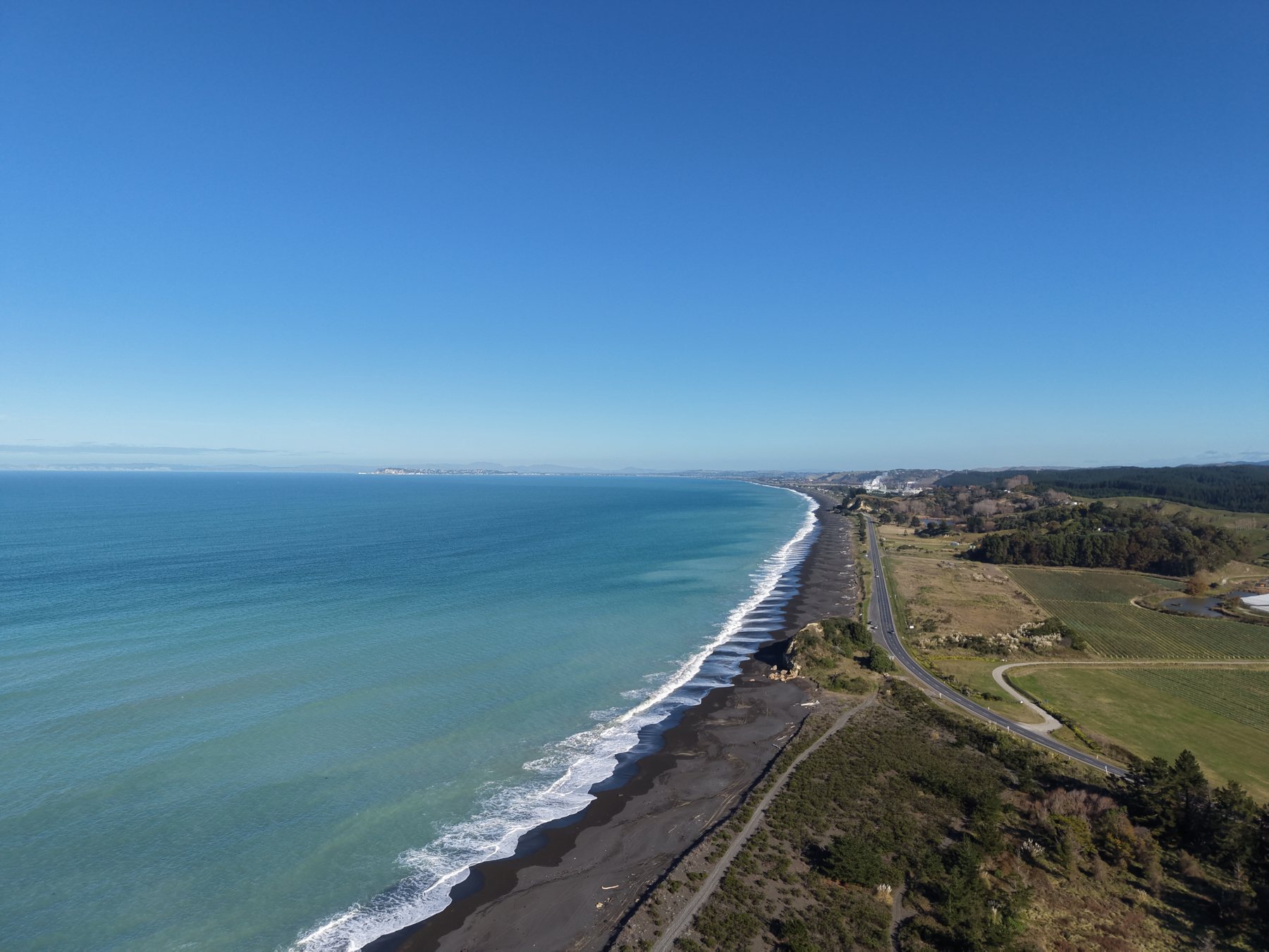 The Hawke’s Bay coast from the air: surf on a black-sand beach, the coast road, orchards and paddocks running inland under a clear sky