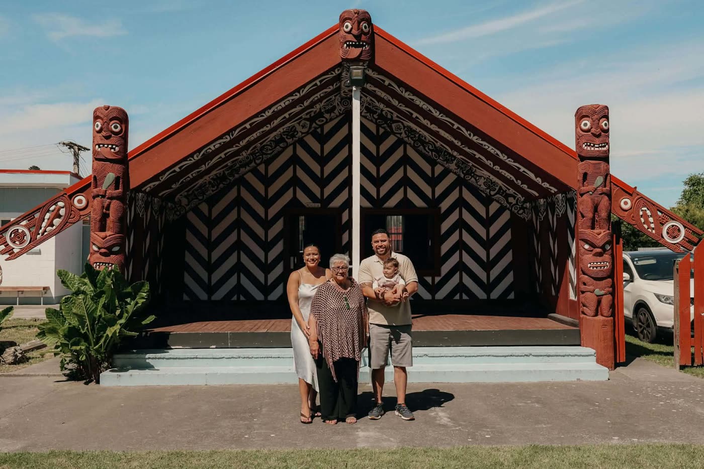 The wharenui — carved tekoteko and maihi framing whānau gathered before it