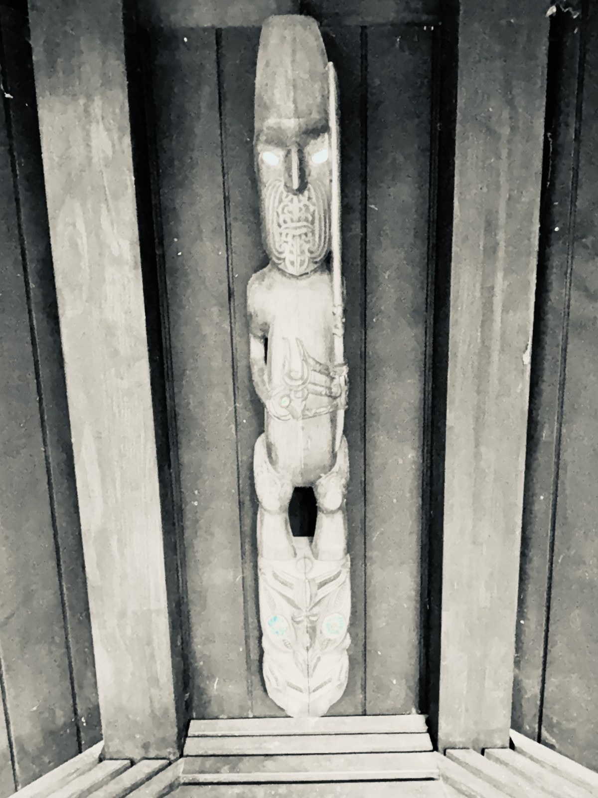 Carved tekoteko standing in the shelter of the waharoa, in black and white, eyes catching the light
